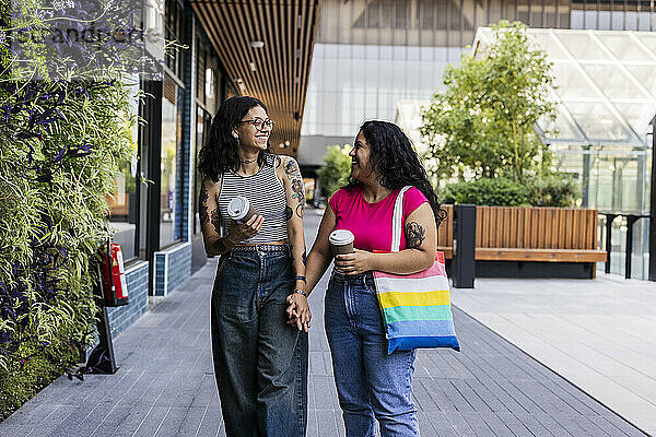 Young gay couple holding disposable cups walking on sidewalk at street