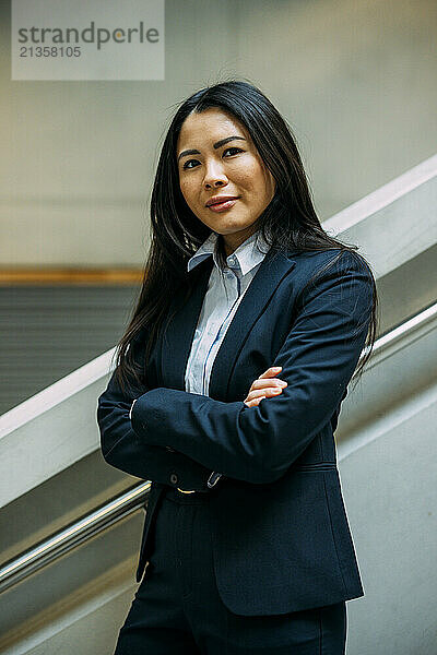 Confident businesswoman with arms crossed standing at staircase at office