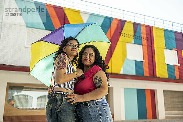 Smiling gay couple standing under multi colored umbrella in front of building