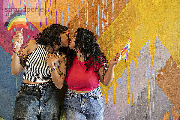 Affectionate lesbian couple holding pride flag and kissing near painted wall