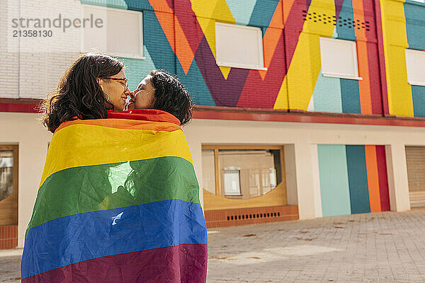 Romantic lesbian couple wrapped in LGBTQIA flag and kissing in front of building