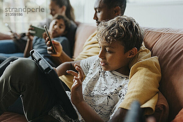 Curly haired boy using tablet PC while sitting with father on sofa at home