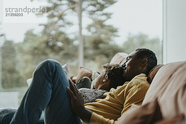 Multiracial boy leaning on father sitting at sofa in living room at home