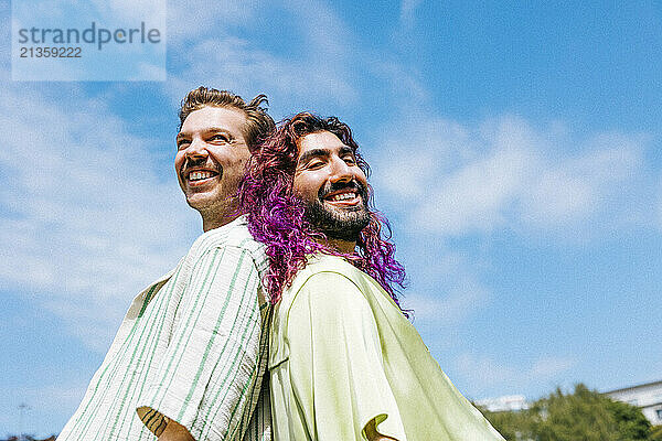 Happy male friends standing back to back under cloudy sky