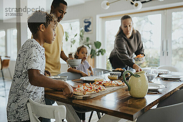 Boy keeping pizza board while parents setting dining table for lunch at home