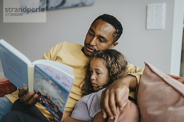 Father reading book with daughter while sitting at home