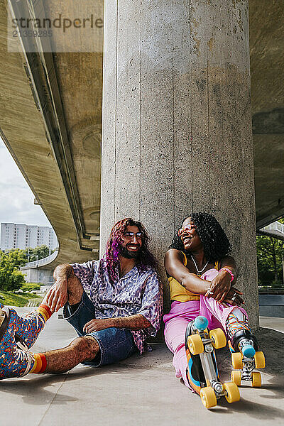 Smiling young woman wearing roller skates sitting with male friend near pillar of bridge