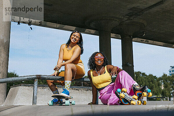 Portrait of happy female friends wearing roller skates sitting near overpass at sunny day