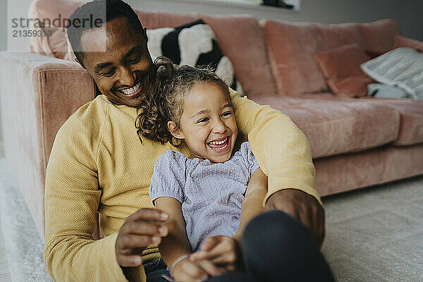 Happy girl playing with father sitting near sofa in living room at home