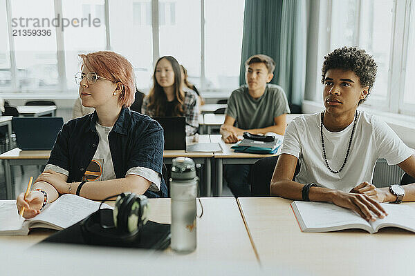 Non-binary person sitting by male friend at desk in classroom