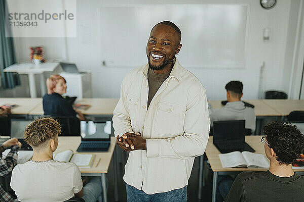 Portrait of happy male teacher standing amidst students sitting at desk in classroom