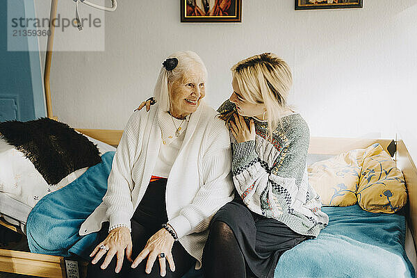 Female caregiver talking with elderly woman sitting on bed at retirement home