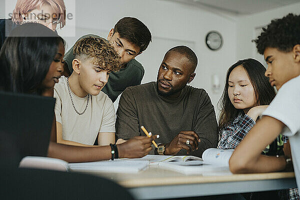 Teenage girl discussing with male teacher while sitting with classmates at desk