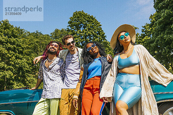 Fashionable male and female friends standing side by side near vintage car at park