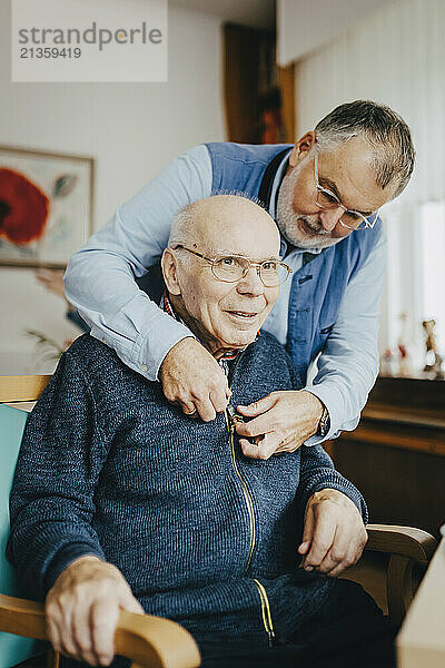 Senior man helping elderly friend in wearing jacket while sitting on chair at retirement home