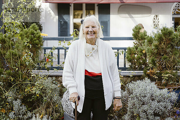 Happy senior woman holding walking cane while standing in front of retirement home