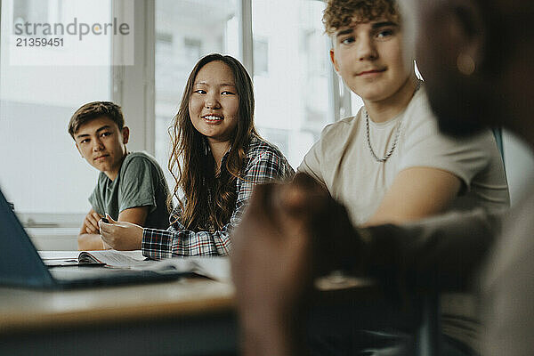 Smiling teenage girl with boys listening to male teacher discussing in classroom