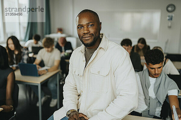 Portrait of smiling male teacher sitting on desk by student in classroom