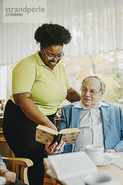 Smiling female caregiver standing by retired man reading book at nursing home