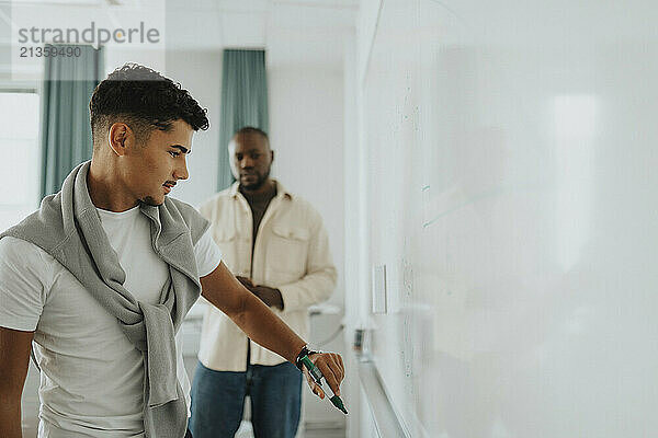 Teenage student with felt tip pen writing on whiteboard with teacher in background