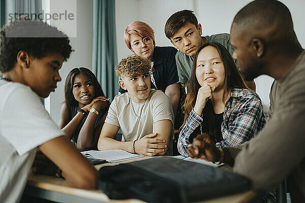 Teenage girls and boys with non-binary person looking at teacher discussing in classroom