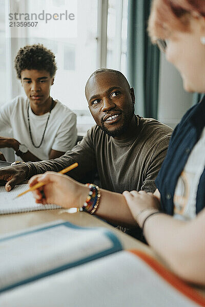 Male teacher discussing while sitting amidst teenage boy and non-binary person in classroom