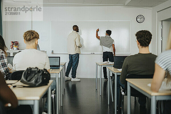 Rear view of male student writing on whiteboard by teacher standing classroom