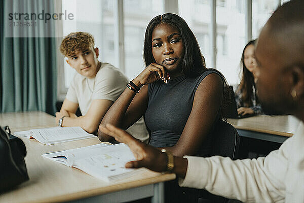 Male teacher gesturing while explaining to teenage boy and girl sitting at desk in classroom