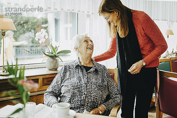 Smiling woman with hand on shoulder of happy senior woman sitting on chair at retirement home