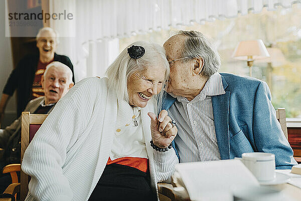 Senior man whispering in female friend's ear while sitting at retirement home