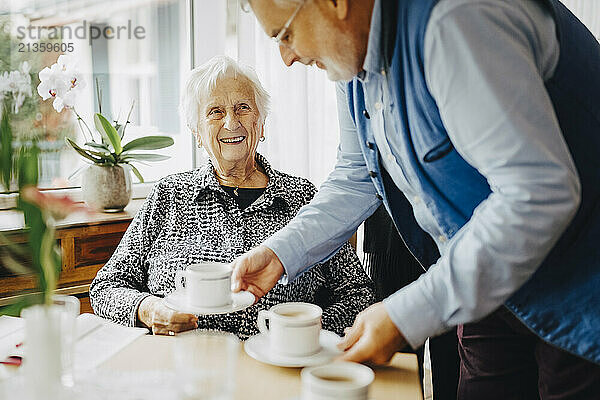 Elderly man serving tea to smiling senior man sitting at retirement home