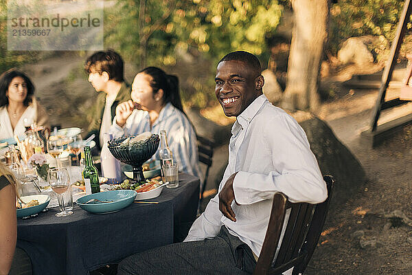 Side view of smiling man sitting on chair with friends at dining table during dinner party