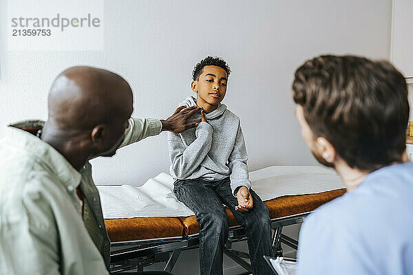 Father explaining neck problem of son sitting on examination table to doctor at clinic