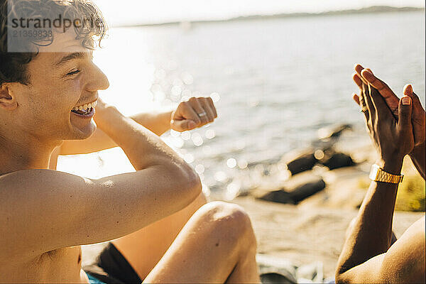 Happy shirtless male friends having fun while sitting near lake at sunny day