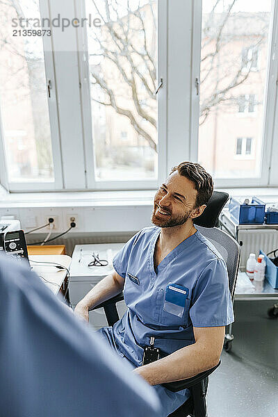 High angle view of male medical expert wearing blue scrubs sitting on chair and talking with coworker in clinic