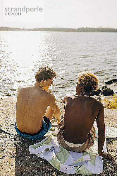 Rear view of shirtless male friends sitting on rock near lake at sunny day