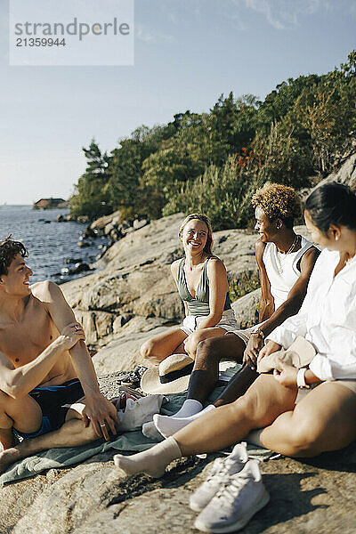 Multiracial male and female friends talking with each other while sitting on rock