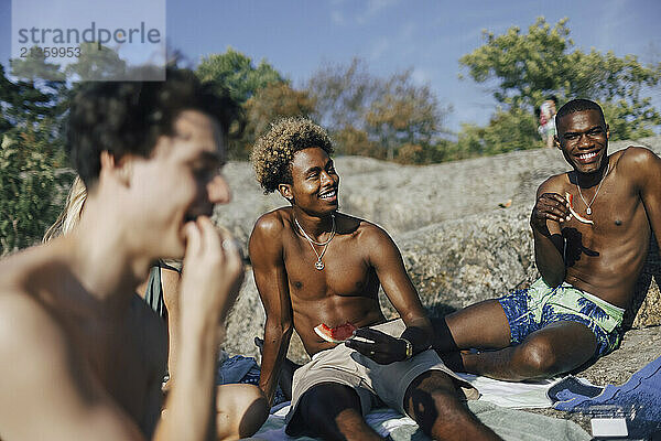 Smiling shirtless male friends eating watermelon while spending weekend with friends