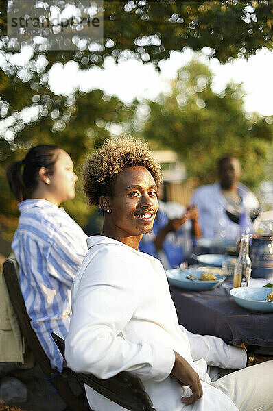 Side view of smiling man sitting on chair with friends during social event in back yard