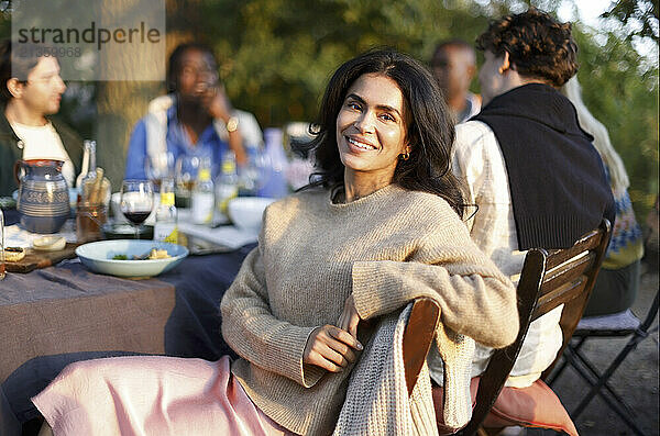 Portrait of smiling woman sitting on chair with friends enjoying during dinner party in back yard