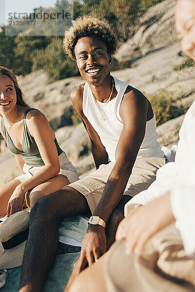 Smiling young man spending leisure time sitting with female friends on rock