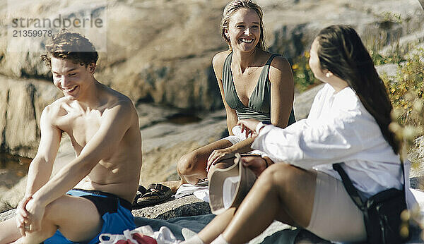 Smiling male and female friends talking with each other while sitting on rock at sunny day