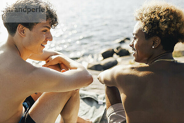 Shirtless young male friends talking with each other while sitting by lake on vacation