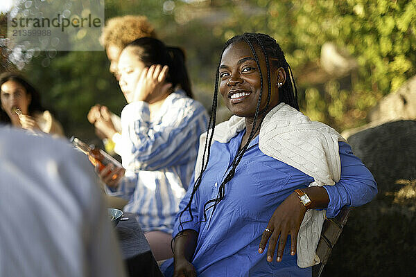 Portrait of smiling woman with braided hair sitting on chair with friends at dinner party in back yard