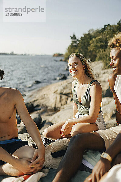 Smiling young woman sitting with friends on rock at sunny day