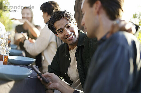 Young man talking with male friend using smart phone while sitting at dinner party in back yard