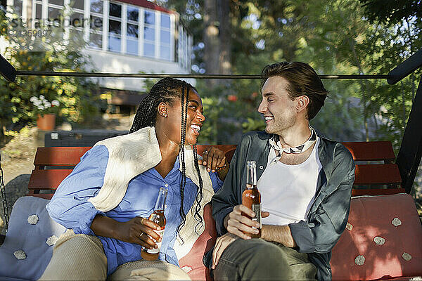 Smiling multiracial male and female holding drink bottles and talking with each other while sitting on bench at party