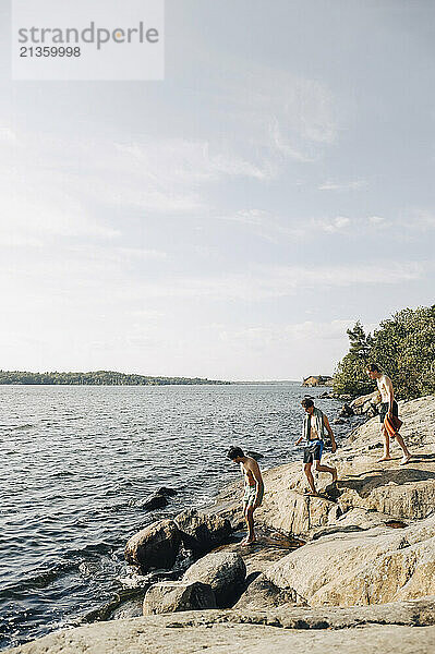 Shirtless male friends moving down on rocks near lake during picnic at sunny day