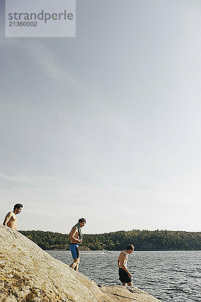 Shirtless male friends walking towards lake against sky at sunny day