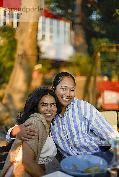 Portrait of smiling woman embracing female friend sitting at dining table during dinner party